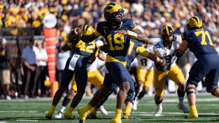 A football player in a navy and yellow uniform with the number 19 throws the ball during a game, surrounded by teammates and opponents on a field with a crowd in the background.
